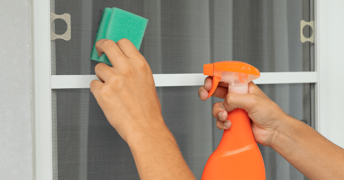 Egy személy zöld szivacsot és narancssárga permetezőt használ a szúnyogháló tisztításához.