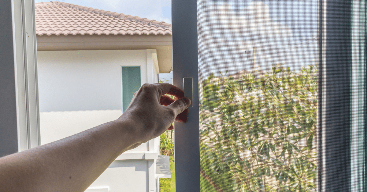 Egy kéz egy ablakot nyit, ahol a szúnyogháló előtt virágzó cserjére látható kilátás nyílik.