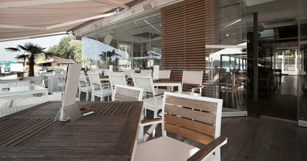 Egy modern étterem terasza, amelyet kitekerhető napellenzővel árnyékoltak. A napellenző teraszra és erkélyre egyaránt ideális megoldás, könnyen feltekerhető és hatékonyan védi a teraszt a napsugárzástól. Az elegáns, fa és fehér székekkel berendezett terasz hangulatos étkezési helyet kínál, ahol a terasz árnyékolás biztosítja a kellemes időtöltést.
