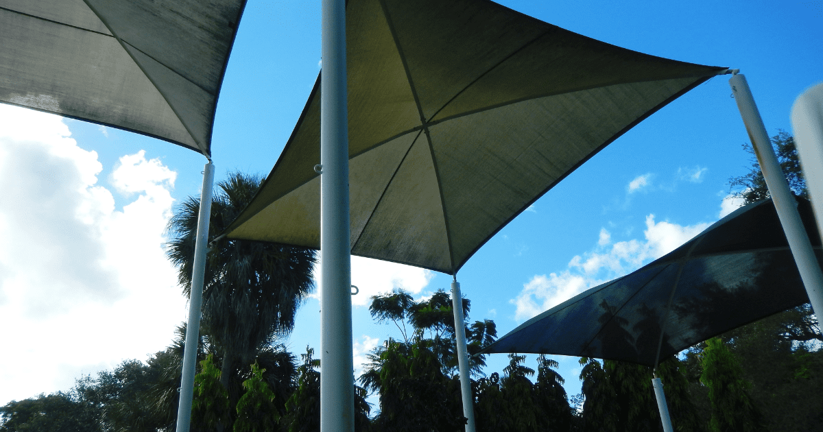 Viharálló terasz árnyékolók egy parkosított területen, magas pálmafák és zöld növények között. Az árnyékolók háromszög alakúak, és masszív oszlopokhoz rögzítve nyújtanak védelmet a napsütés és a szél ellen.